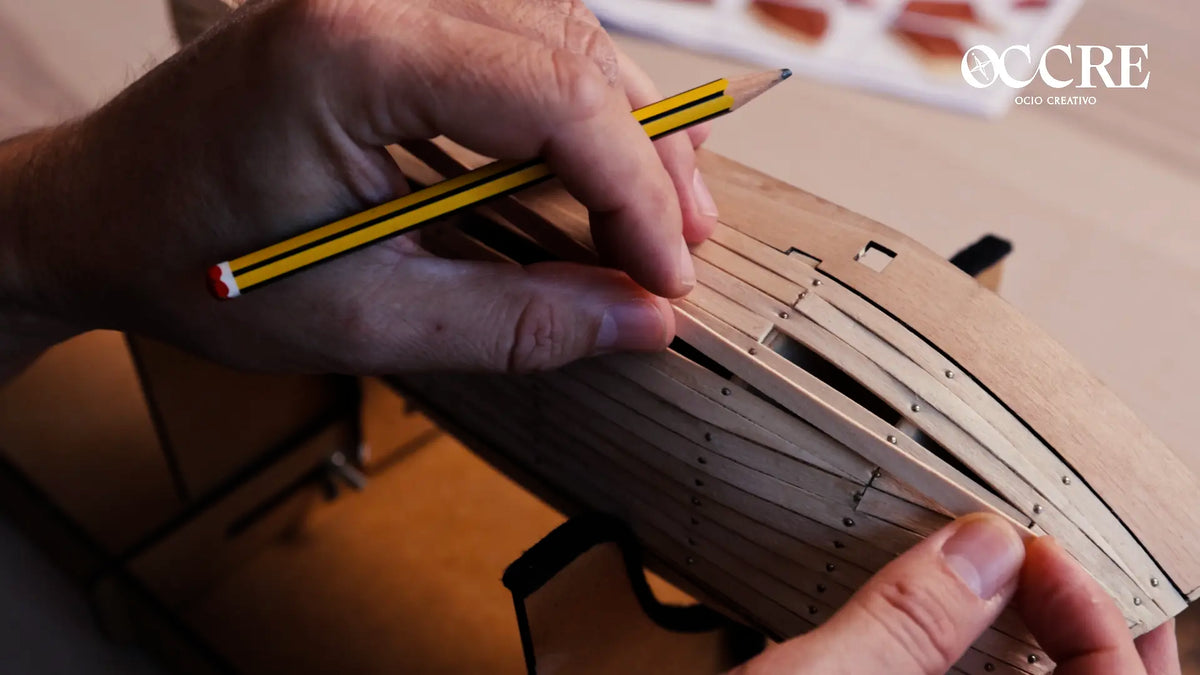 Hands carefully fitting a wooden plank to a model ship hull